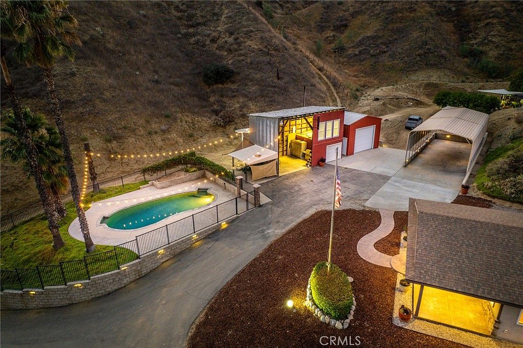 Fenced Pebble Tec pool lit at dusk with string lights, barn and carport behind